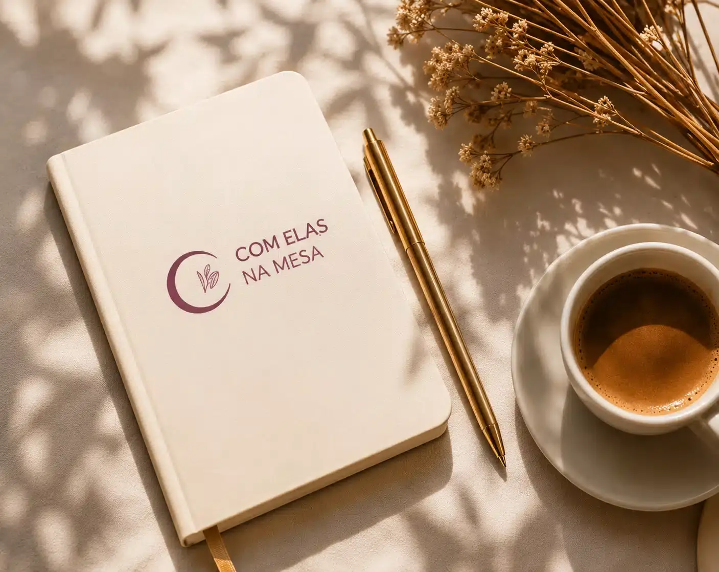 Caderno, caneta e café com a identidade visual Com Elas na Mesa